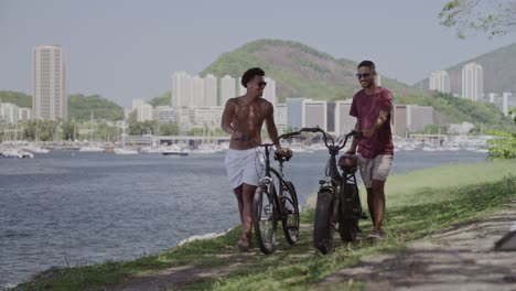Two Men Walking Bicycles Along a Scenic Waterfront with a Cityscape Background