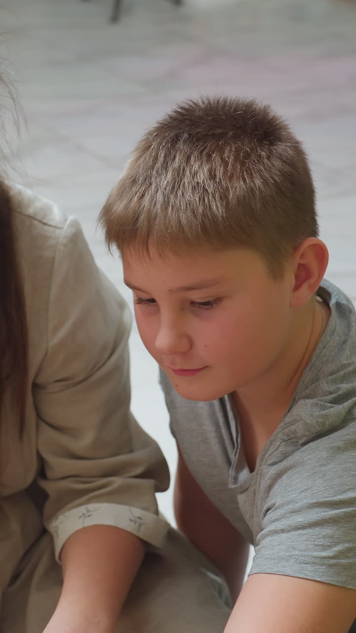 niño caucásico estudiando de cerca una bola de nieve, sentado en el suelo de baldosas junto a su padre, mirada concentrada y manos cuidadosas, sutiles luces bokeh y borde de árbol, camiseta gris casual, momento de tradición colaborativo