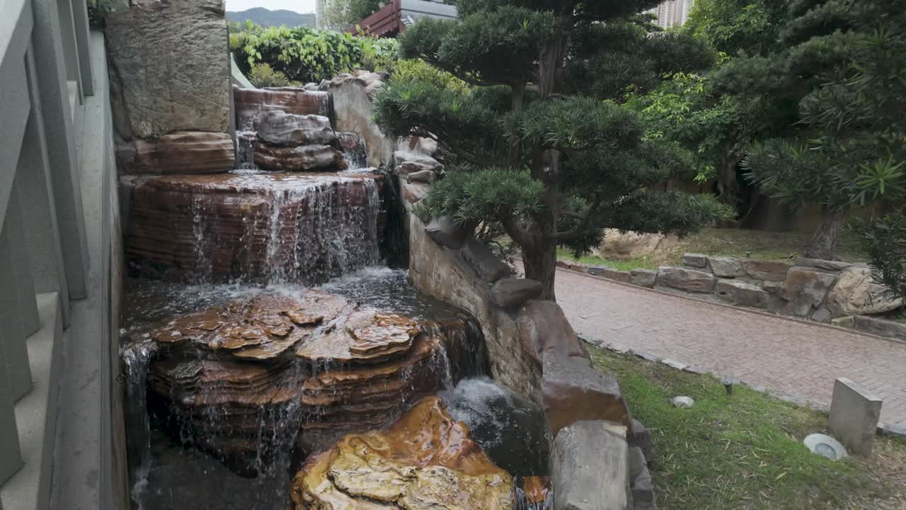 Cascata artificiale di roccia che scende a cascata su gradini di pietra in uno stagno circondato da alberi verdi nel tranquillo Nan Lian Garden a Hong Kong, giornata di sole