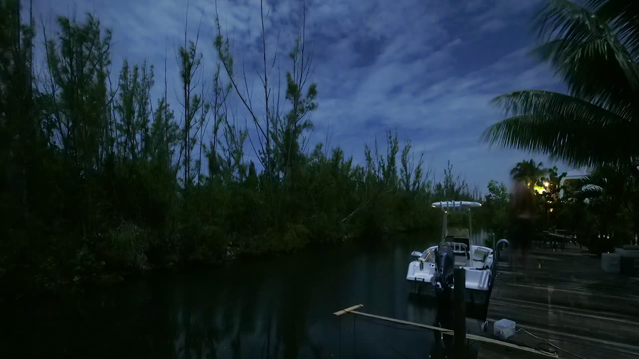 Nuvens noturnas passam rapidamente sobre um canal de quintal durante uma lua cheia. Um homem aparece no quadro pescando com uma lanterna de cabeça no final.