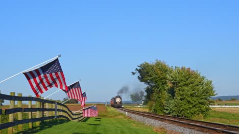 a view of a line of gently waving american flags on a fence by farmlands as a steam passenger train blowing smoke approaches during the golden hour