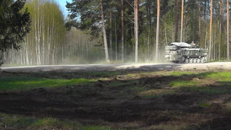 british warrior fv510 armored personel carrier tank driving fast on a gravel road with dust cloud behind it and armed soldiers on top of it in a forest during a sunny day with pine trees in foreground
