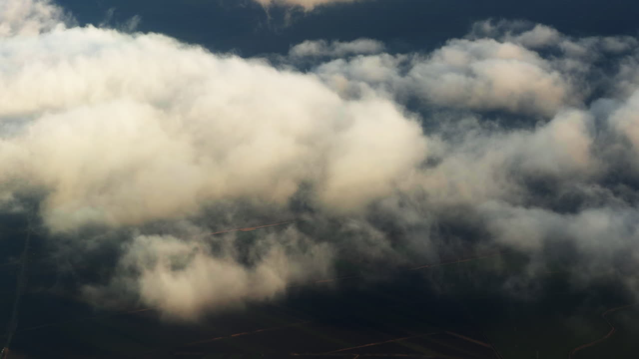 Luftfoto af dramatiske skyer, der driver hen over landskabet og afslører marker og snoede veje nedenunder, fanger et atmosfærisk vejrbillede fra et flyveperspektiv i stor højde.