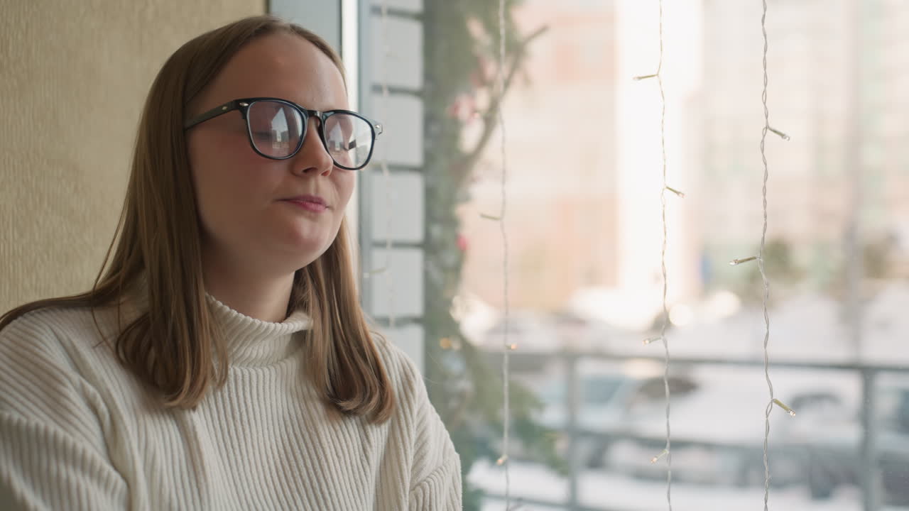 happy woman in glasses holds spoon with chocolate cake in cozy cafe with decorative lights and snowy street view through window creating warm cheerful mood