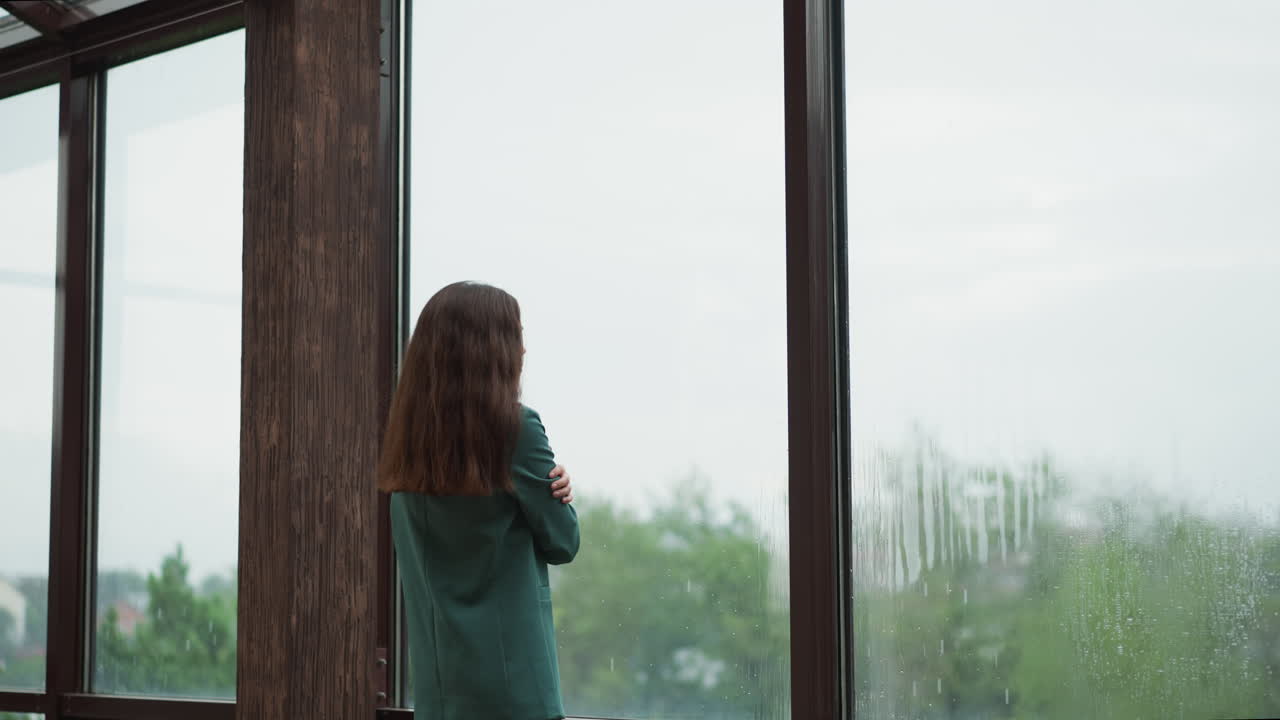 여자는 유리 위로 떨어지는 물방울을 쳐다보고, 사업가 여성은 테라스에서 기업 세계의 혼란에서 휴식의 순간을 찾습니다.