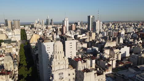 Aerial-lowering-on-Barolo-Palace-tower-in-tree-lined-May-Avenue-surrounded-by-Buenos-Aires-buildings-at-golden-hour