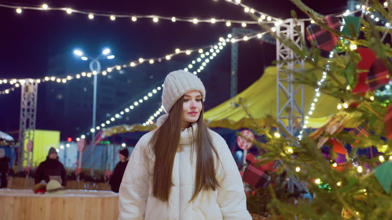 Young woman in cozy winter attire approaches a beautifully decorated Christmas tree, admiring and smelling the fresh pine branches, soft bokeh background with glowing festive lights