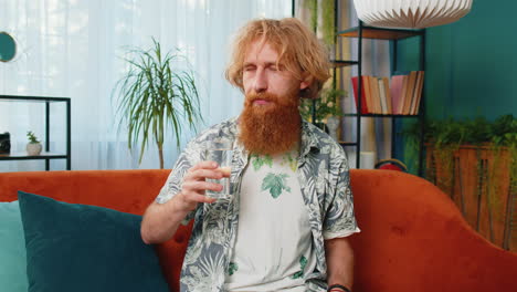 Thirsty-Caucasian-man-sits-at-home-holding-glass-of-natural-aqua-make-sips-drinking-filtered-water