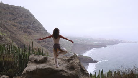 Slow-motion:-Woman-meditates-in-yoga-warrior-pose-by-ocean,-beach,-and-rock-mountains,-epitomizing-motivation,-inspiration,-and-a-healthy-lifestyle's-harmony-with-nature's-fitness-concept