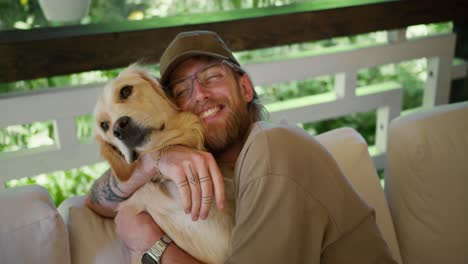 a blond guy with a beard and glasses in a light brown t-shirt and a cap strokes a large light-colored dog on a sofa in a gazebo in nature