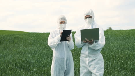 caucasian researchers in protective suit using tablet and laptop while doing pest control in the green field