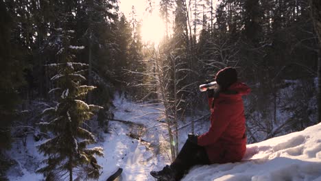 girl in red coat sitting on cliff edge drinking tea on a winter sunny day - medium shot, slow motion