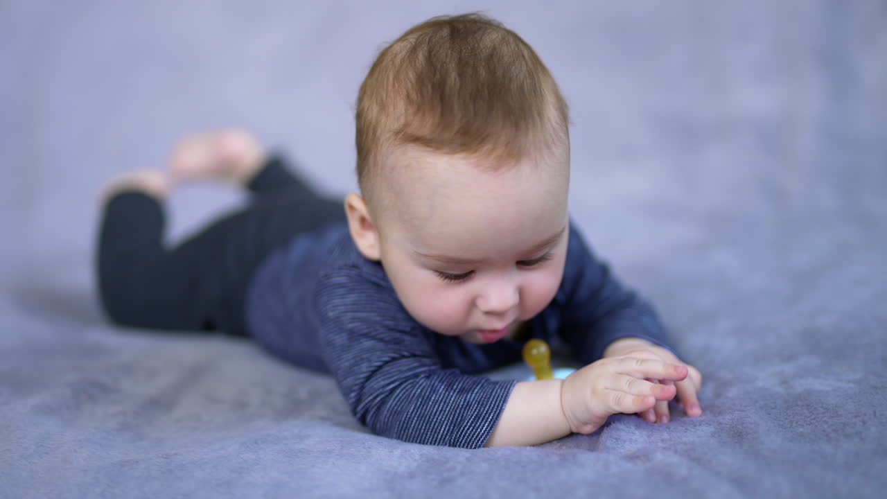 Lovely toddler in blue clothes lying peacefully on the bed. Cute child looks at pacifier with interest and then around himself.