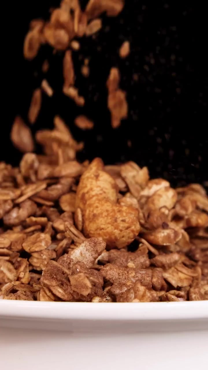 Fiocchi di granola cadono a cascata in una ciotola bianca, su uno sfondo nero, creando un'immagine dinamica e materica