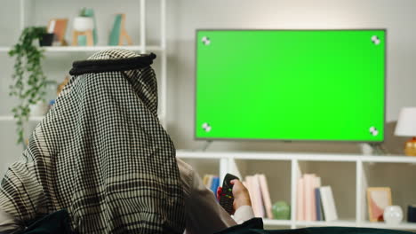 Middle eastern man watching tv with chroma key close-up. Person watches television with green screen in living room. Wearing traditional Islamic clothes