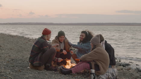 group of teenage friends sitting around the bonfire while they roasting sausages and talking on the seashore