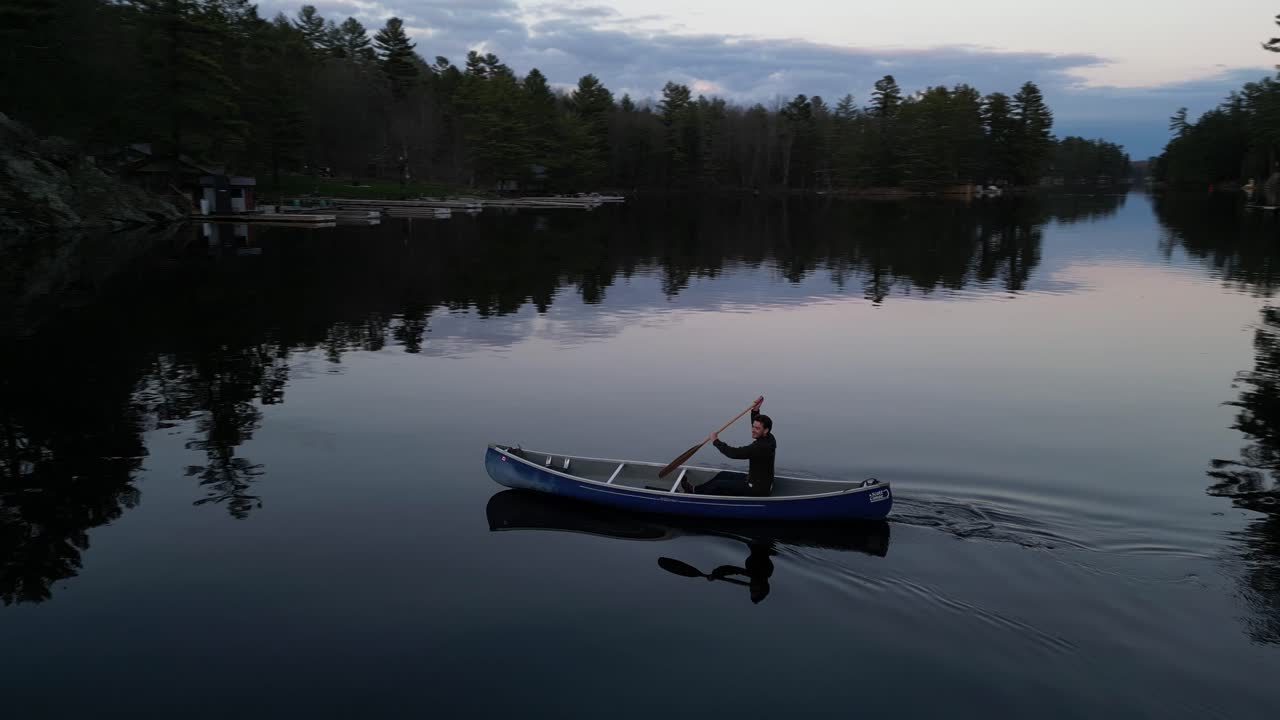 Person canoeing on a serene lake at twilight, with reflections of trees on the calm water and a peaceful atmosphere