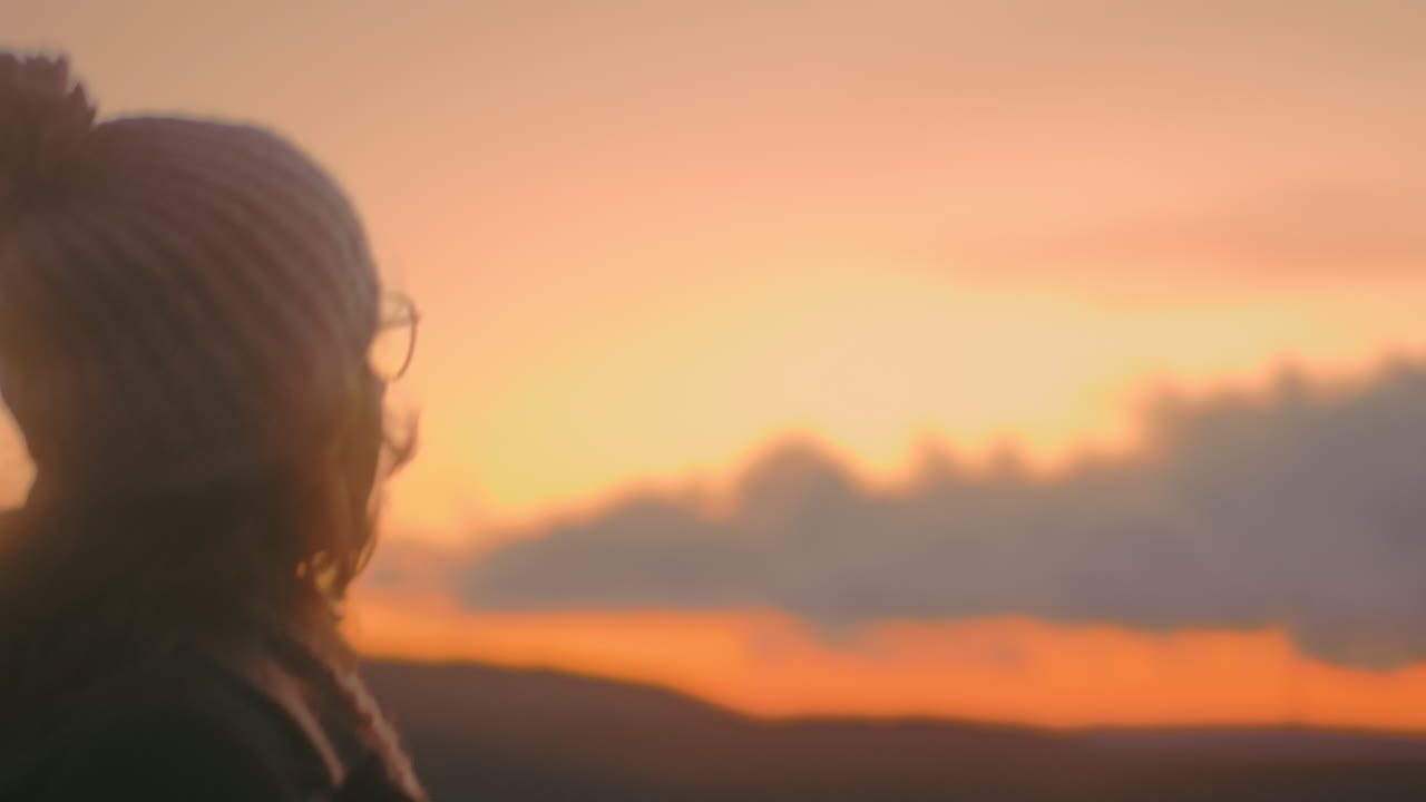 Millennial woman in gray coat walks towards working wind turbines in Portugal on a beautiful day. Camera follows her from behind, highlighting renewable energy, sustainability, and nature's serenity.