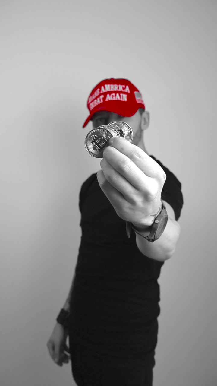 Grayscale footage of a man with tattoos and a red MAGA Make America Great Again hat lifting up tree Bitcoin cryptocurrency shiny and reflective coins in front of the camera showing them in close up.