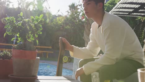 Asian-male-teenager-sitting-with-skateboard-on-sunny-day-in-garden