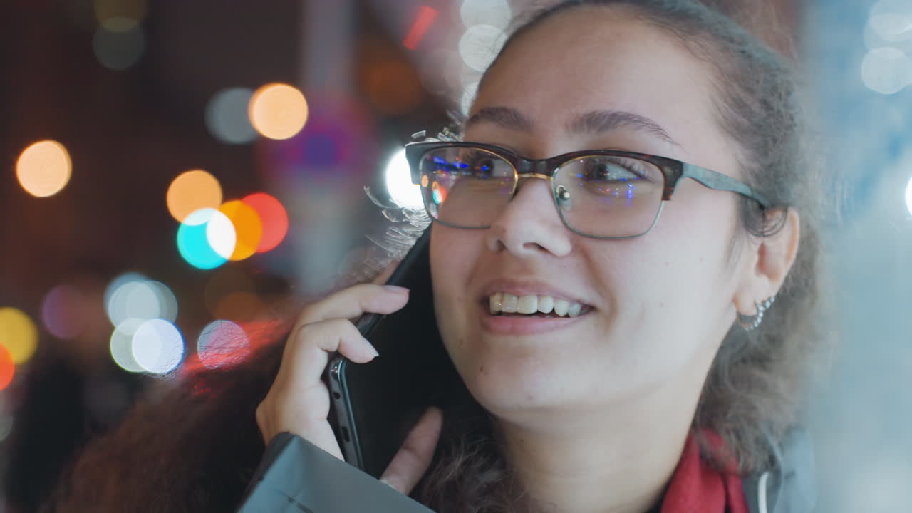 Young woman talking on phone night, glasses, colorful bokeh lights, urban street background, candid closeup showing shifting expressions from smile to surprise, winter coat and scarf, handheld mobile