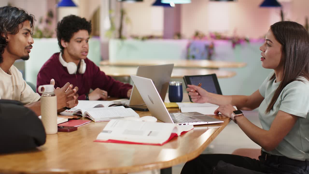 Diverse students around wooden table discussing coursework using laptops after center man gesturing