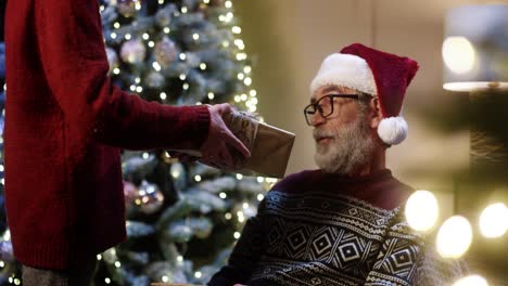 Close Up Of Loving Nice Grandson Giving Christmas Gift Box To Old Happy Grandpa In Santa Hat While Sitting In Decorated Room Near Glowing Tree And Hugging Him