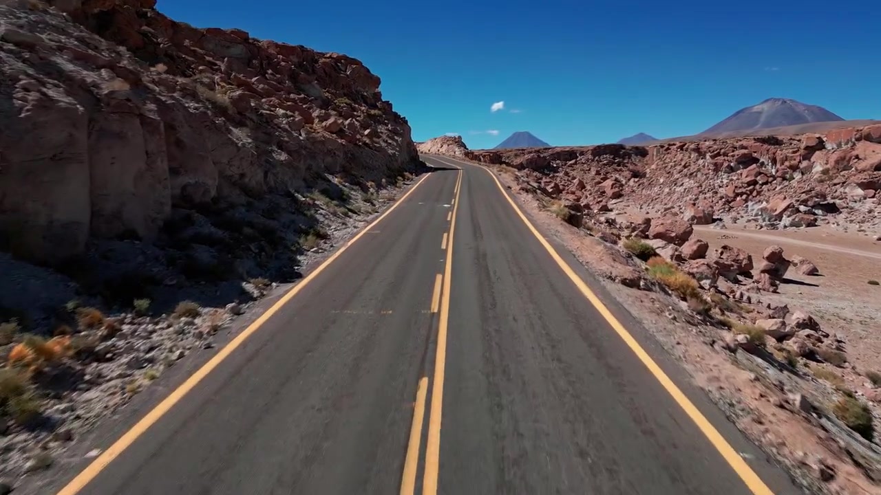 Vuelo rápido y bajo hacia adelante a lo largo de una carretera desértica pavimentada que atraviesa un cañón de roca roja con picos volcánicos visibles al frente bajo un cielo azul claro de altiplano