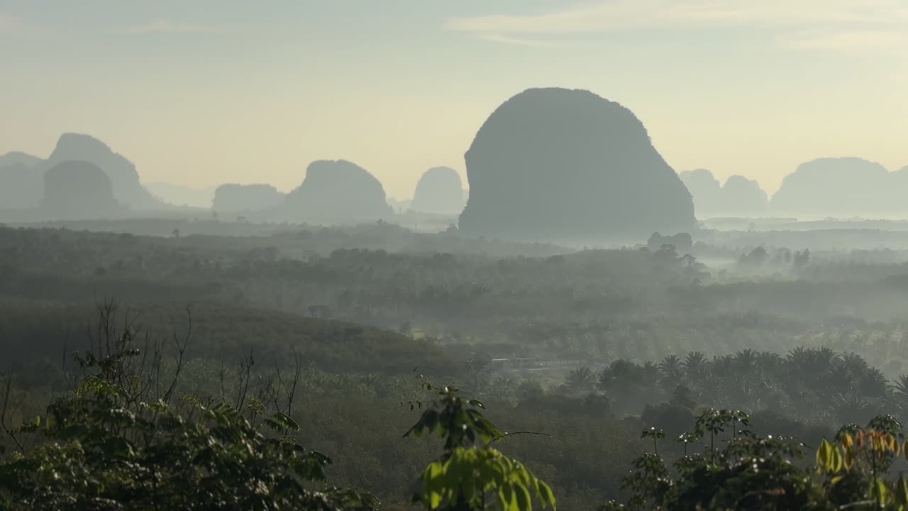 Valle nebbiosa all'alba con lontane sagome calcaree e una foresta stratificata a Din Daeng Doi, Krabi, Thailandia, che mostra una leggera foschia, luce calda e un paesaggio sereno.
