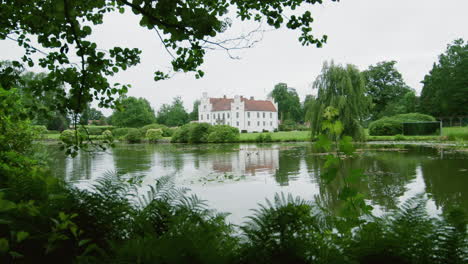 Aufschlussreiche-Aufnahme-Eines-Alten-Magischen-Feenschlosses-Aus-Baumeiche-In-Der-Nähe-Eines-Wunderschönen-Sees-Mit-Malerischen-Blumen-Und-Klarem-Wasser-An-Einem-Bewölkten-Tag.-Romantischer-Ritterpalast-Im-Nordischen-Skandinavien