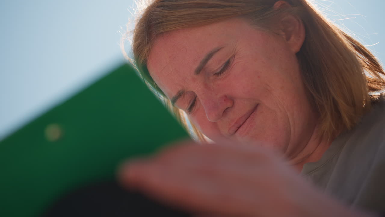 Close up of woman looking down with soft smile in bright sunlight, warm natural glow highlighting face and hair, evoking emotion, calm reflection, and peaceful outdoor atmosphere under clear blue sky