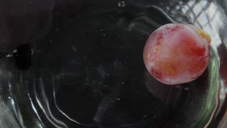 a single grape falls into a glass of water, creating ripples and bubbles in slow motion