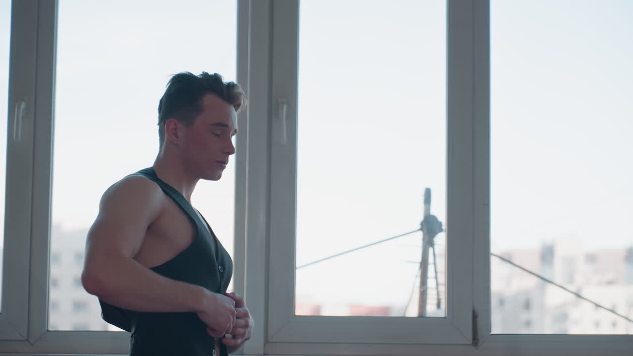 stylish muscular man stands near tall apartment window gently fastening black sleeveless vest while morning city skyline glows outside; soft sunlight outlines tattoo on chest