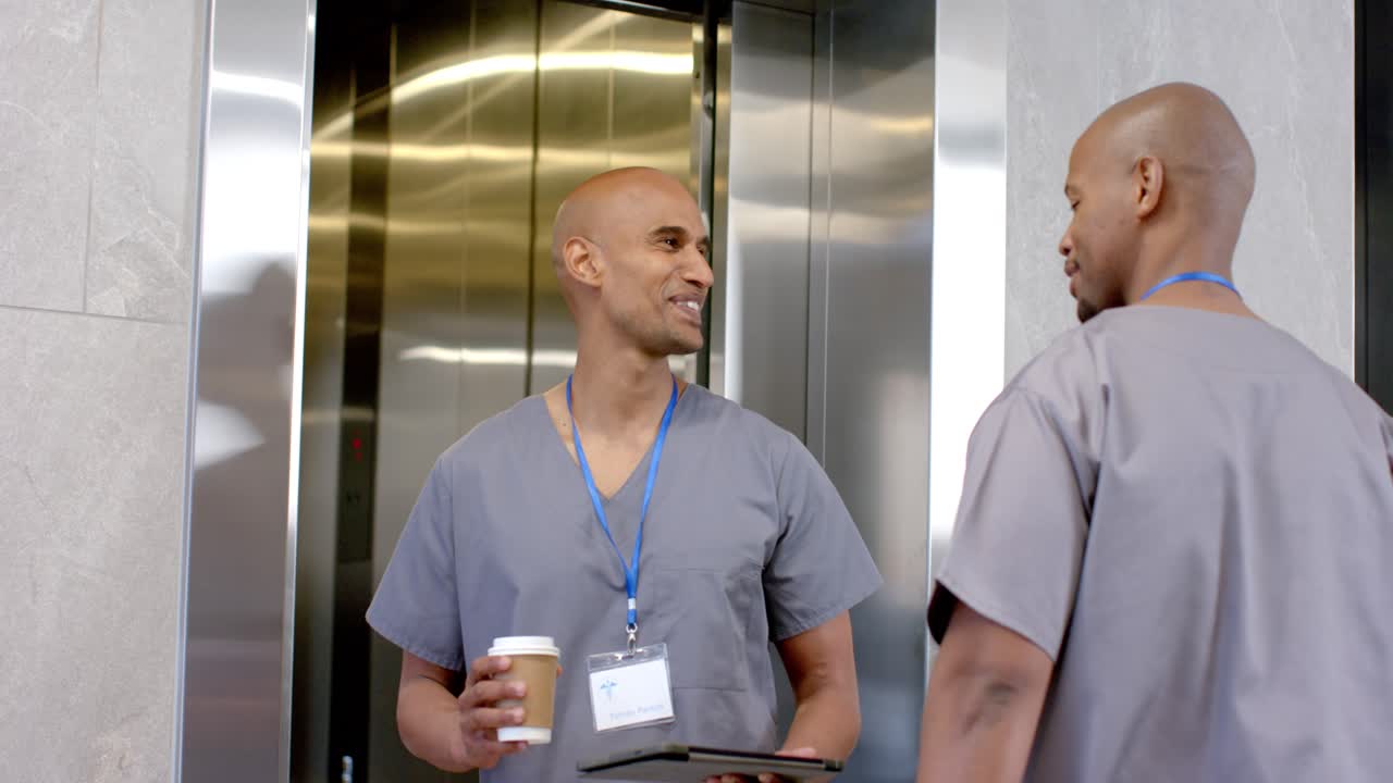 2 African American male staff exiting elevator greeting in clinic hall, holding coffee tablet phone