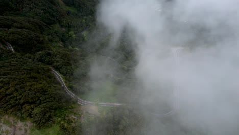 aerial view over a curvy mountain road, nestled among green plants and framed by thick-white clouds, la gomera island