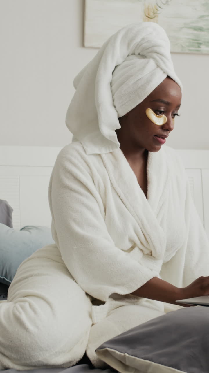 Black Girl Using Laptop Wearing under Eyes Patches and White Bathrobe at Home