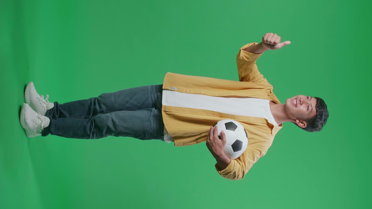 녹색 스크린 배경에서 축구를 응원하는 동안 엄지손가락을 나타내는 공을 가진 아시아 소년의 전체 몸의 측면 시각