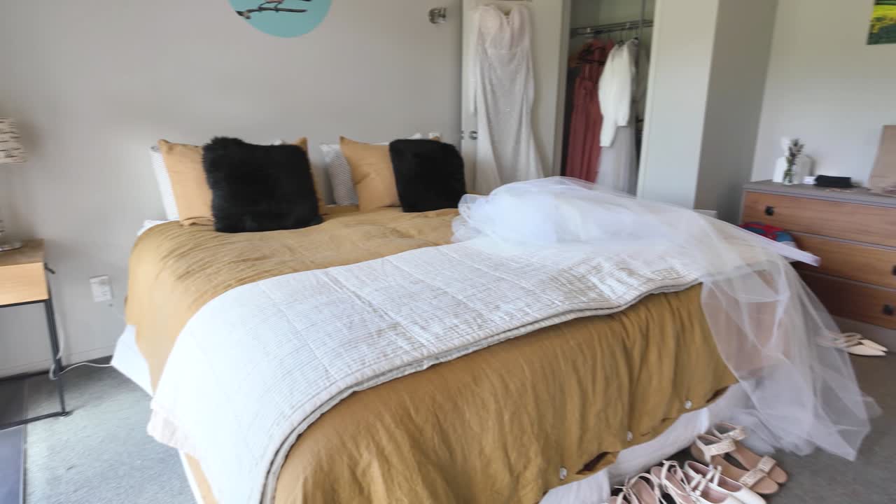Una semplice scena di una camera da letto con una camicia bianca appesa a una porta di vetro, un letto con coperte dai toni caldi e una finestra che si affaccia su un campo