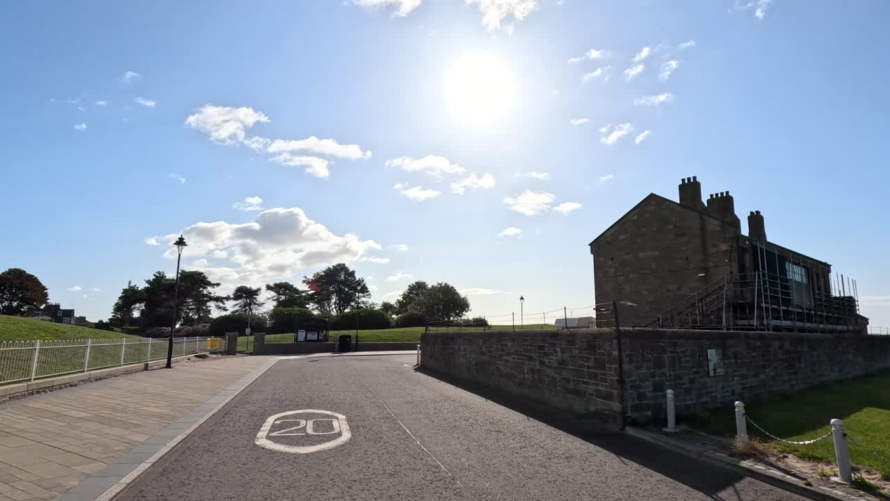 Camera moves smoothly along a quiet street beside a historic stone castle and green lawns under bright morning sunlight in Broughty Ferry, Dundee