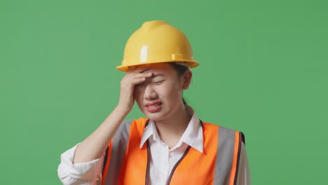 안전 헬을 입은 아시아 여성 엔지니어가 녹색 스크린 배경 스튜디오에서 일하는 동안 두통을 고 있습니다.
