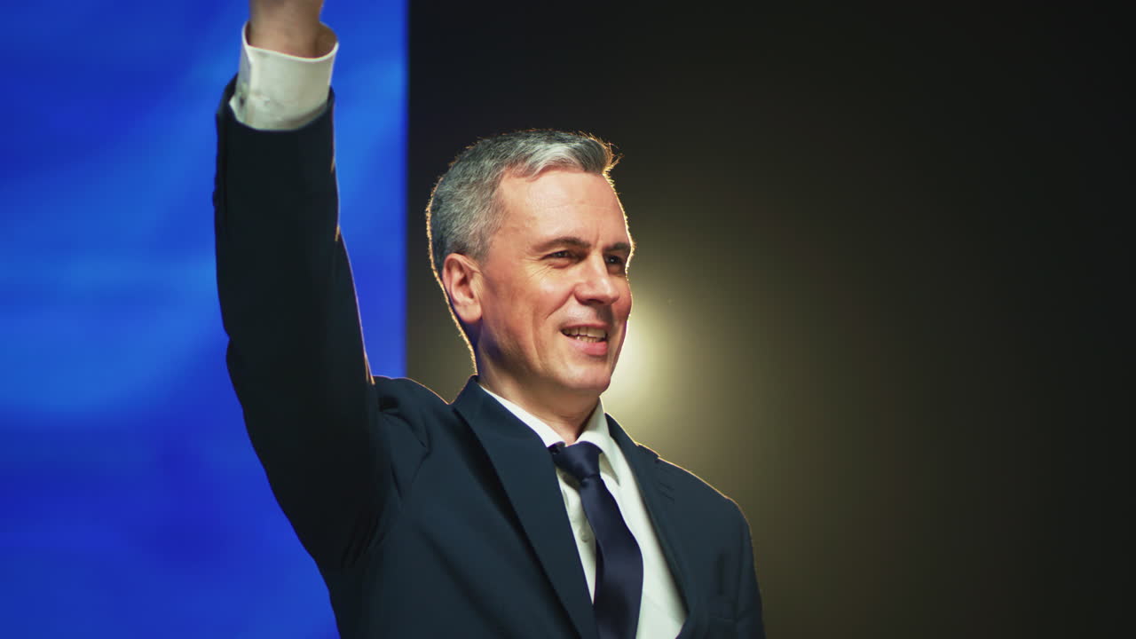 Mature Politician Waving Hand from Stage Zoom out View of Friendly Middle Aged Man in Suit Waving Hand and Clasping Hands over Head while Greeting Audience from Stage against European Flag