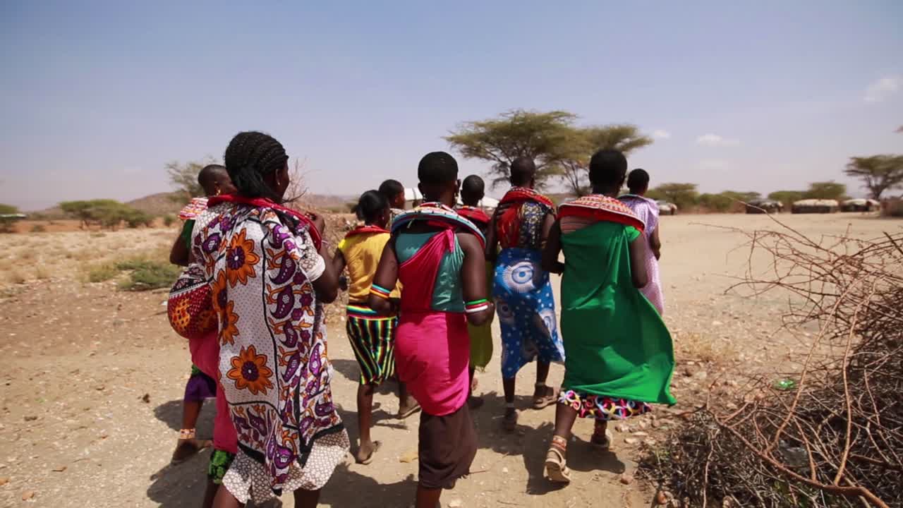 POV walking along with Maasai local tribe in traditional colorful clothing through small village in the drylands of Maasai Mara National Reserve in Narok, African Savanna, Kenya, Africa.
