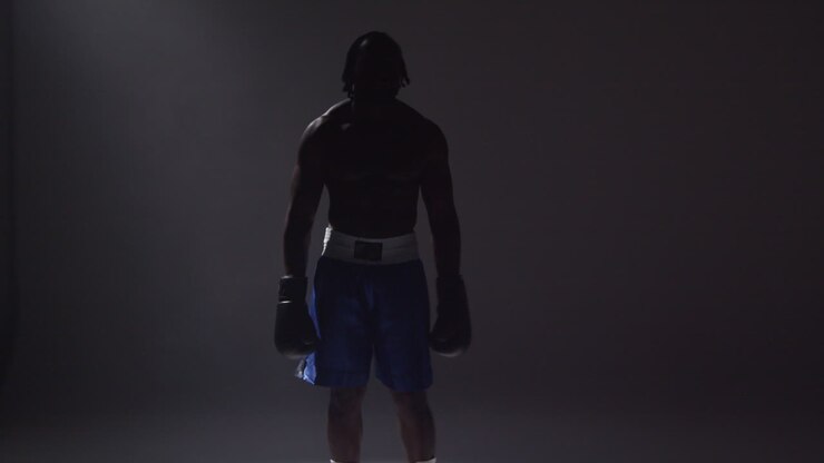 Toma de estudio en silueta con iluminación de clave baja de un boxeador con guantes de pie en un haz de luz sobre un fondo gris 1