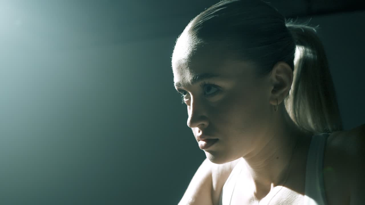 Closeup from the side on a woman’s face and shoulders as she trains, with the camera gliding to the front. Strong backlight creates dramatic highlights, capturing intensity and focus mid-workout