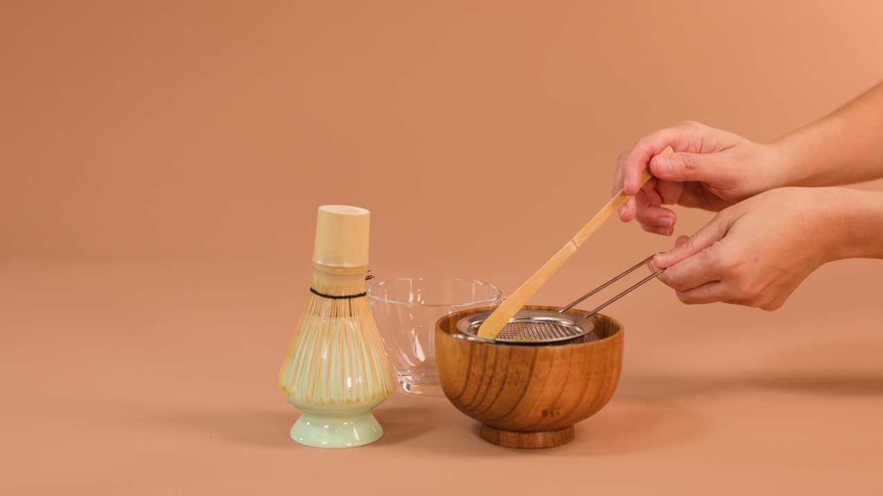 Hender sikter matcha-pulver i en treskål ved hjelp av bambusvisp og sil, myk studiolyssetting