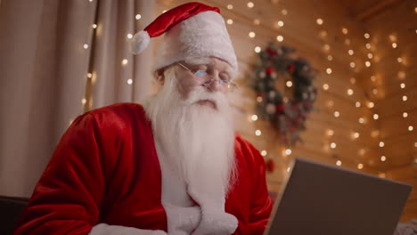 close-up of santa claus working remotely from home sitting on the couch with a laptop near the christmas tree working with a laptop