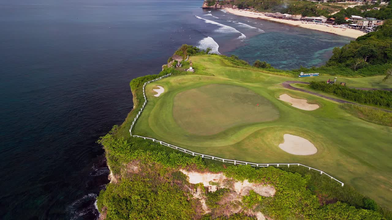 Top-down aerial of seaside golf resort showing green fairways, sand bunkers, paved paths, steep coastal cliffs, clear shallow reef water and white tropical beach under bright daylight