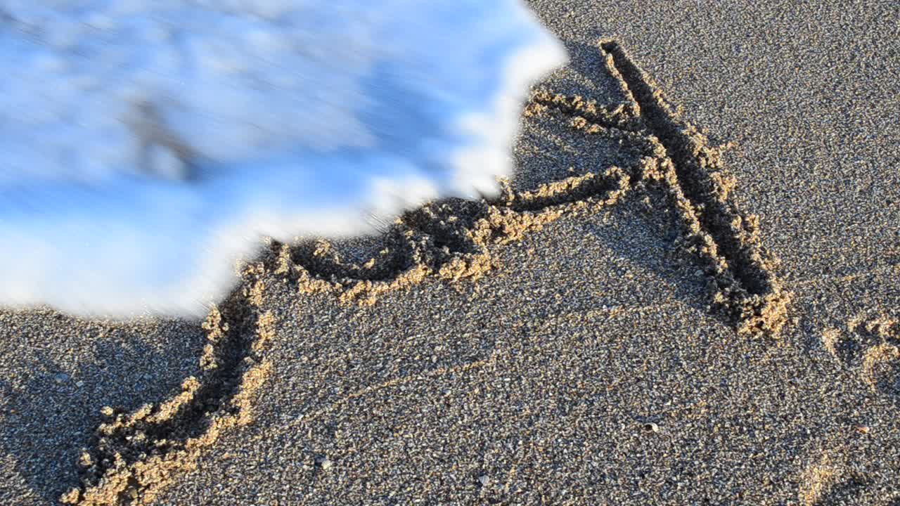 Inskripsjon juli på sand. Inskripsjonen vaskes bort av en havbølge. Filming på stranden.