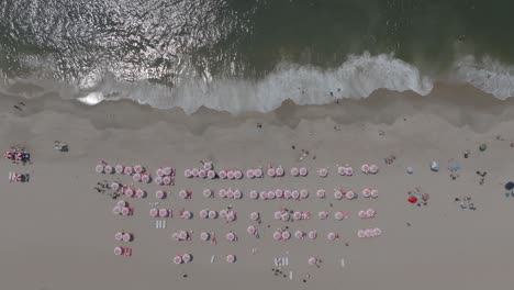 Toma-De-Arriba-Hacia-Abajo-De-Un-Día-Perfecto-En-La-Playa-Con-Sombrillas-Rosas-Uniformes-Y-Las-Olas-Rompiendo-En-La-Playa.