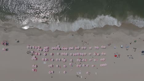 disparo de arriba hacia abajo de un día de playa perfecto con paraguas rosas uniformes con las olas chocando en la playa